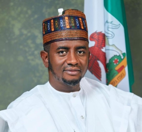 Senator Shehu Buba Umar in white traditional attire in front of the Nigerian coat of arms flag